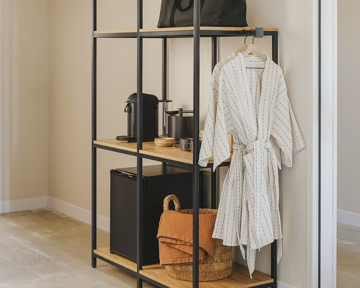 Open concept hotel wardrobe with black metal frame, wooden shelves, Nespresso coffee maker, striped robe, and woven storage