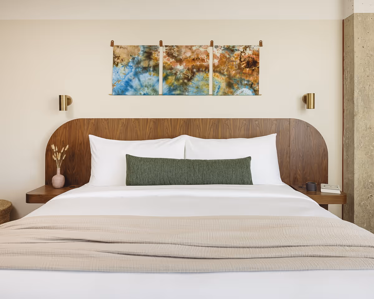 Modern hotel bedroom with curved walnut headboard, white bedding, green bolster pillow, and colorful tie-dye wall art