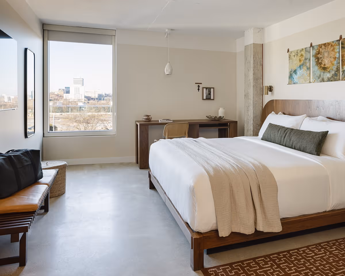 Modern hotel room with king bed, wooden platform frame, concrete floors, large window with city view, and minimalist decor.