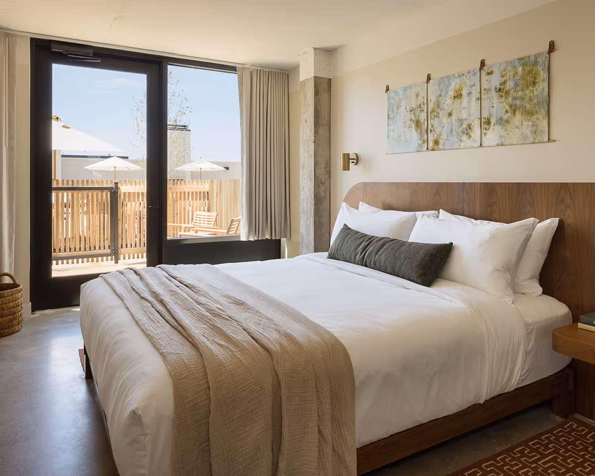 Modern hotel bedroom with king bed featuring white linens, wooden headboard, and balcony access with outdoor seating area.