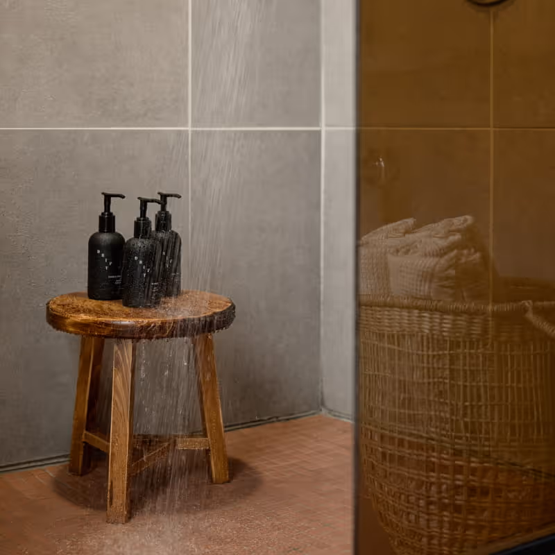 A wooden stool holds three black Drift pump bottles in a running shower, with towels in a wicker basket nearby.