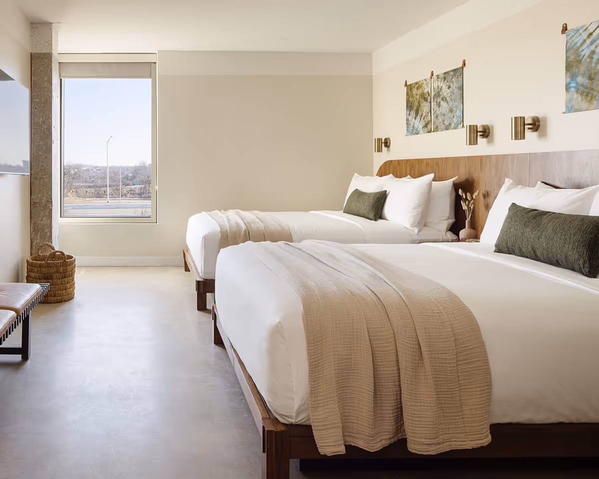 Modern hotel room with two queen beds featuring white linens, olive green accent pillows, wooden headboard, and polished