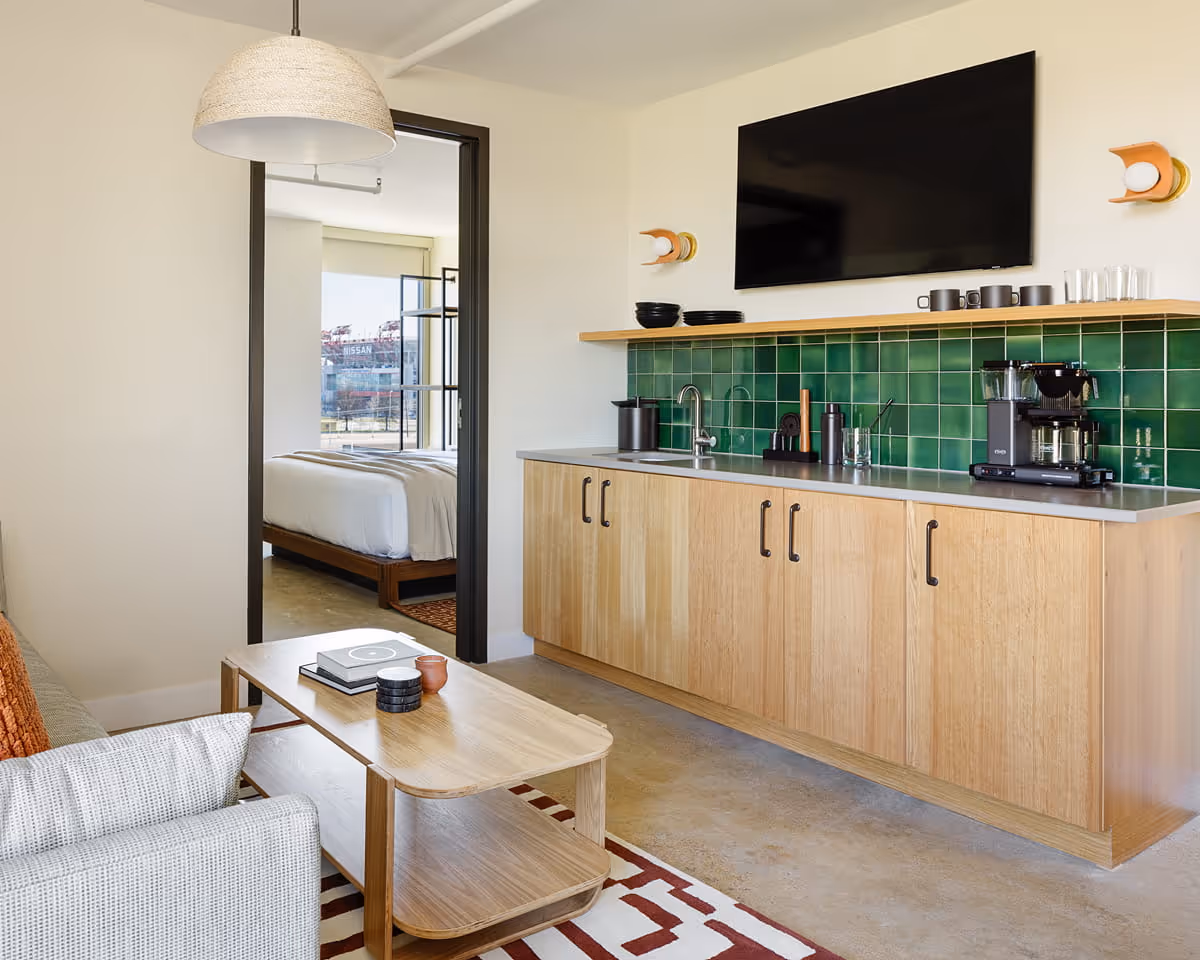 Hotel suite living area with kitchenette featuring green tile backsplash, light wood cabinets, wall-mounted TV, and view