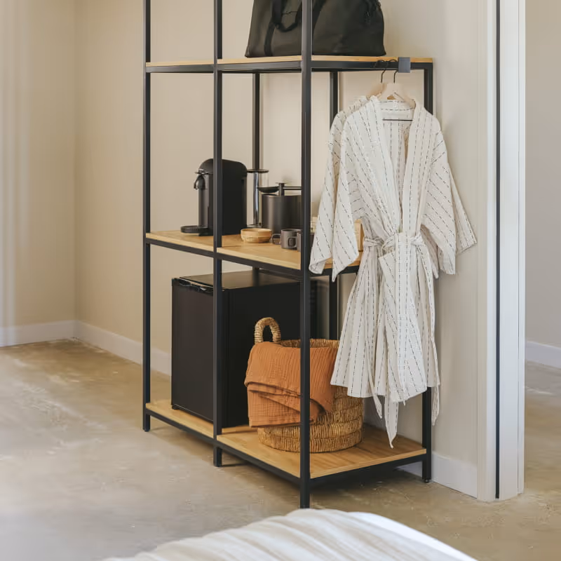 Open shelving unit with black metal frame displaying robe, coffee maker, mini fridge, woven basket, and travel bag in hotel