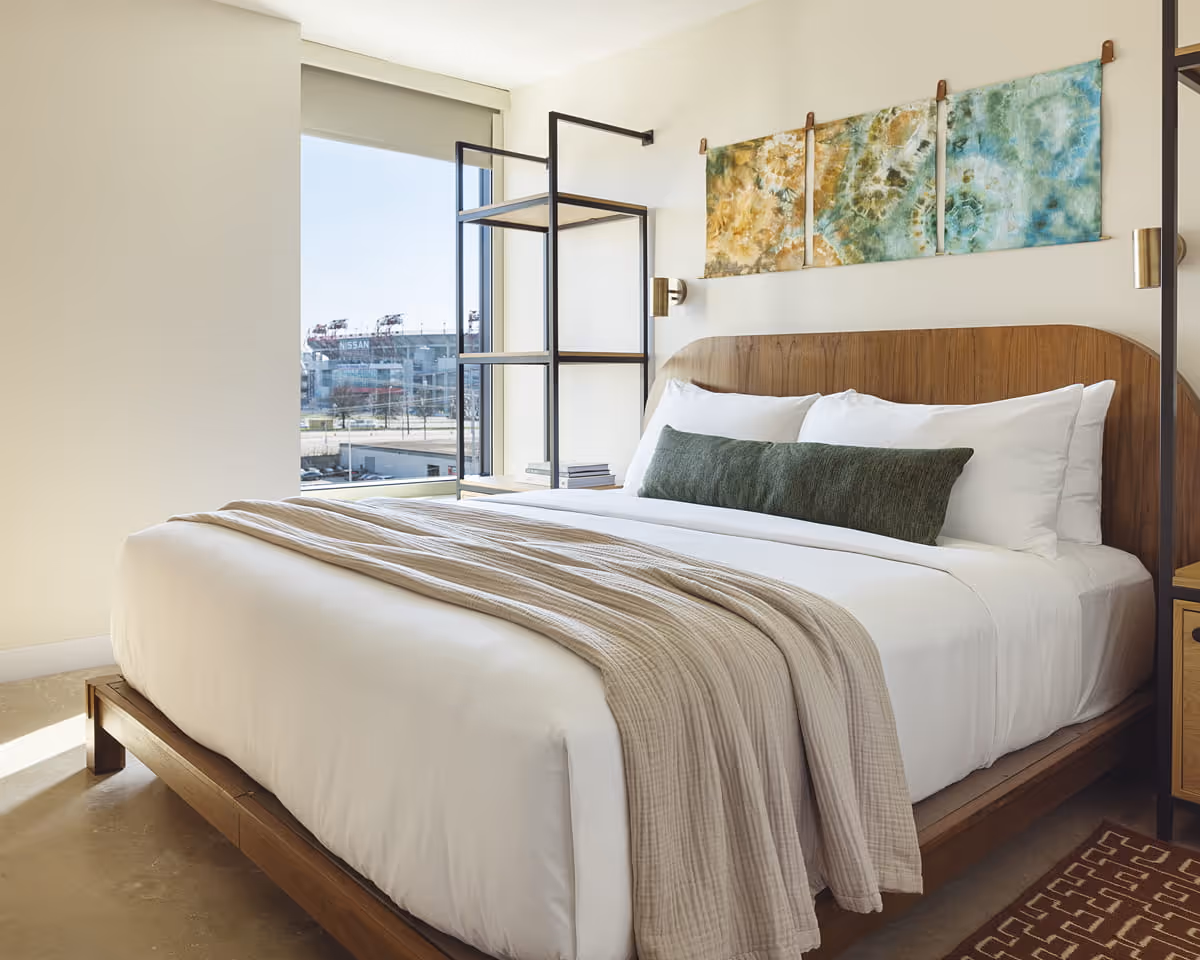 Modern hotel bedroom with wooden headboard, white bedding, green accent pillow, and tie-dye artwork with Nissan Stadium view