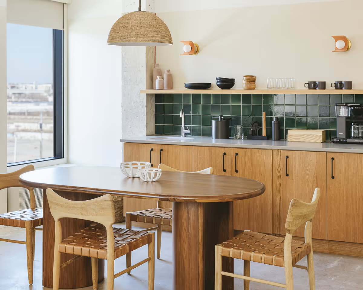 Modern kitchen with green tile backsplash, oak cabinets, oval walnut dining table, and woven leather chairs under a rattan