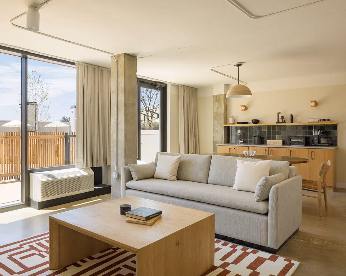 Contemporary hotel suite living area with gray sofa, wooden coffee table, geometric rug, kitchenette, and floor-to-ceiling