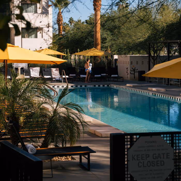 Hotel pool area with yellow umbrellas, lounge chairs, palm trees, and a guest walking poolside on a sunny day.