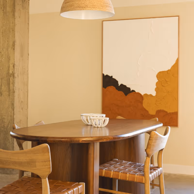 Warm dining area with oval walnut table, woven leather chairs, and abstract earth-toned artwork on a cream wall.