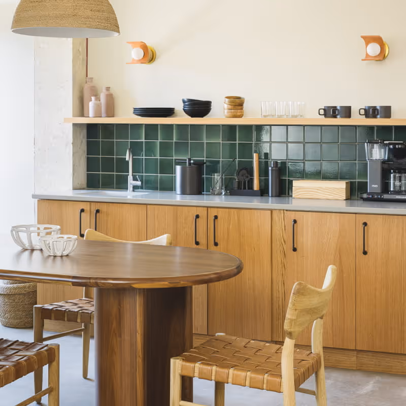 Modern kitchen with green tile backsplash, oak cabinets, wooden dining table, and woven leather chairs in warm tones.