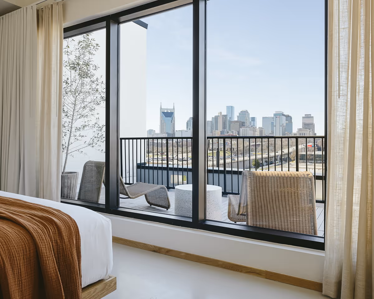 Modern hotel bedroom with floor-to-ceiling windows, balcony seating, and Nashville skyline view featuring the AT&T Building.
