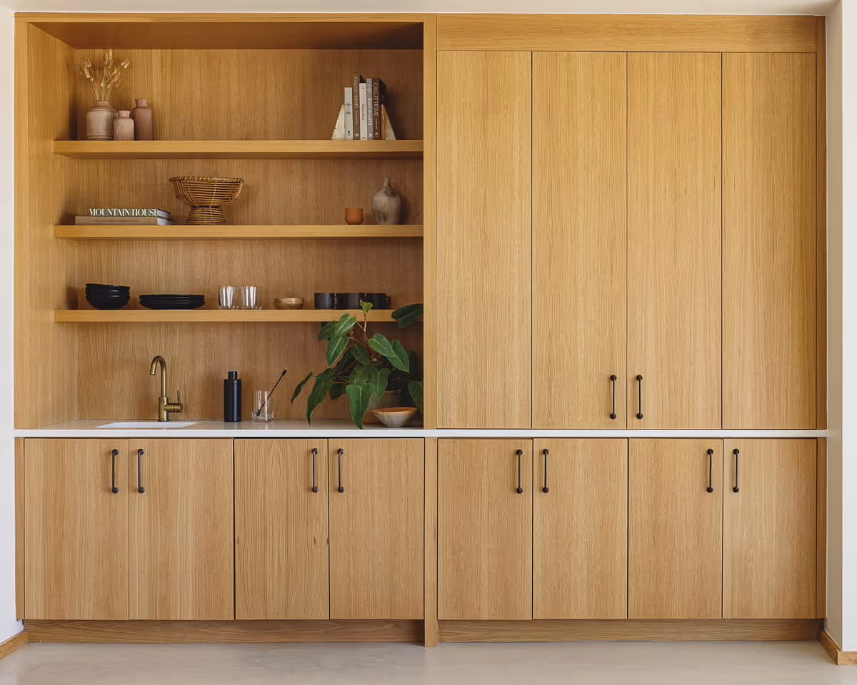 Built-in oak wood cabinetry with open shelving displaying books, ceramics, glassware, and a potted plant above a small wet