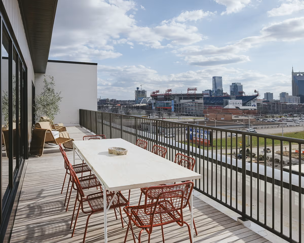 Modern balcony with red metal dining chairs and white table overlooking Nissan Stadium and Nashville skyline on a sunny day.