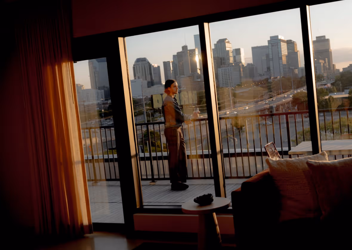 A person stands on a hotel balcony at dusk, gazing at a city skyline with modern high-rise buildings.