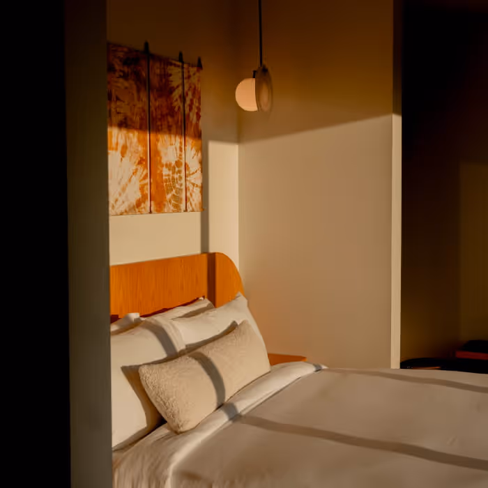 Cozy bedroom with white bedding, plush pillows, curved wooden headboard, and tie-dye wall art illuminated by warm pendant