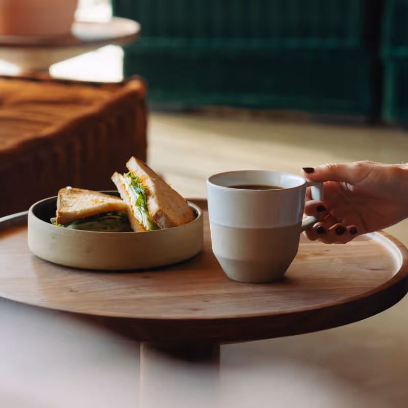 A hand with dark nail polish reaches for a ceramic coffee cup beside a sandwich in a bowl on a round wooden table.