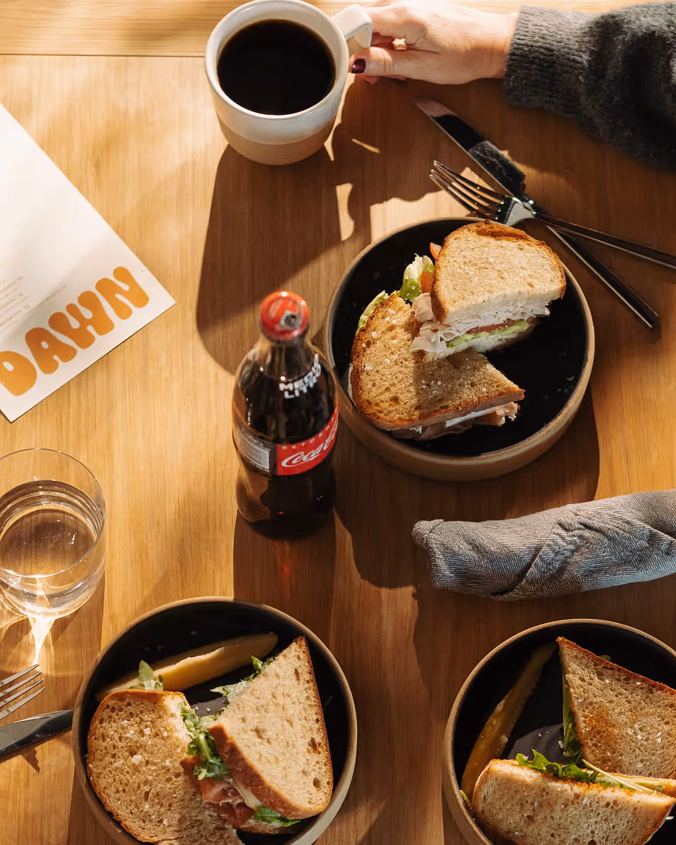 Overhead view of sandwiches on dark plates with a Coca-Cola bottle, black coffee, and a Dawn cafe menu on a wooden table.