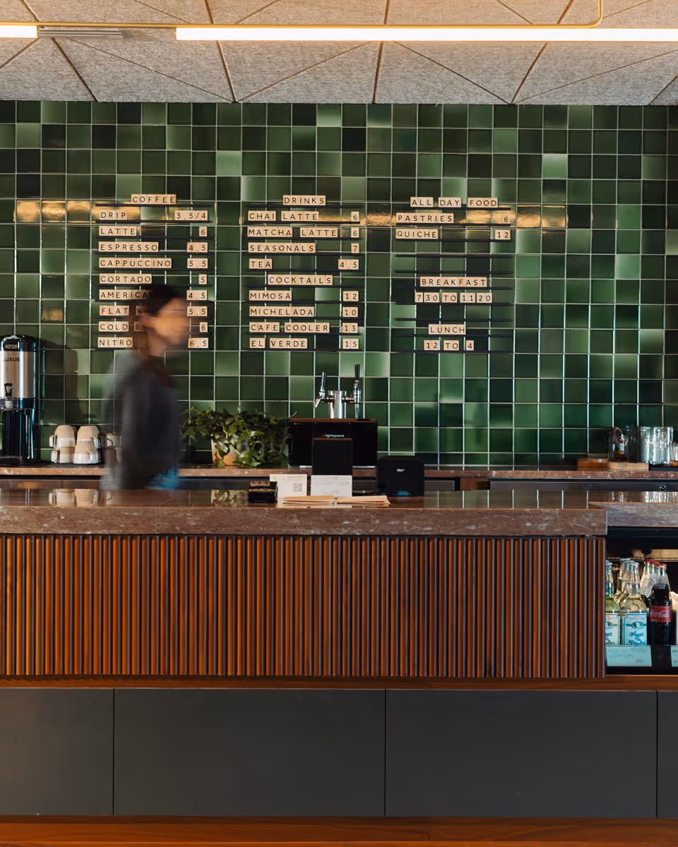 Cafe counter with green tile menu board listing coffee, drinks, and cocktails, with a barista walking behind the counter.