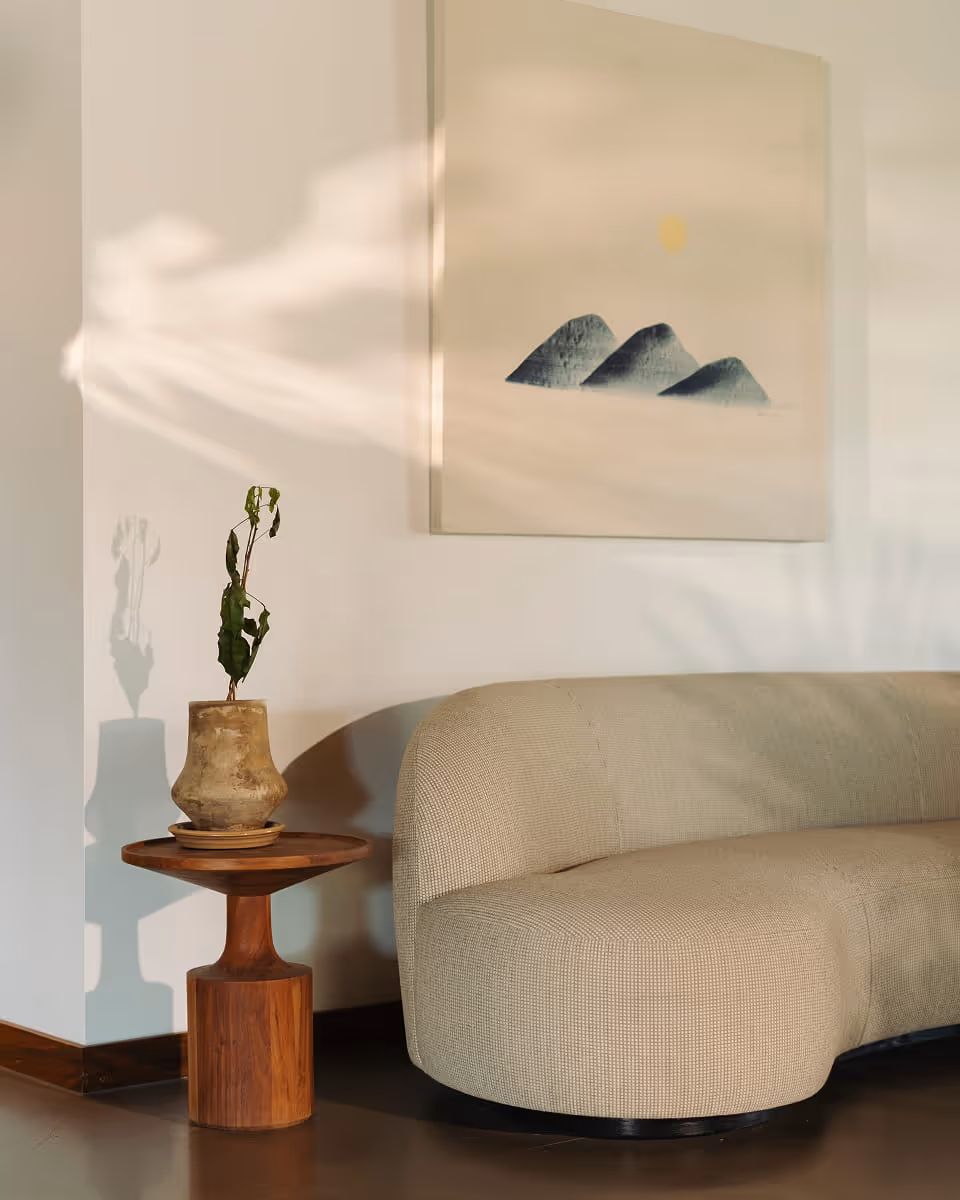 Minimalist cafe lounge corner featuring a curved beige sofa, wooden pedestal side table with a plant, and mountain artwork.