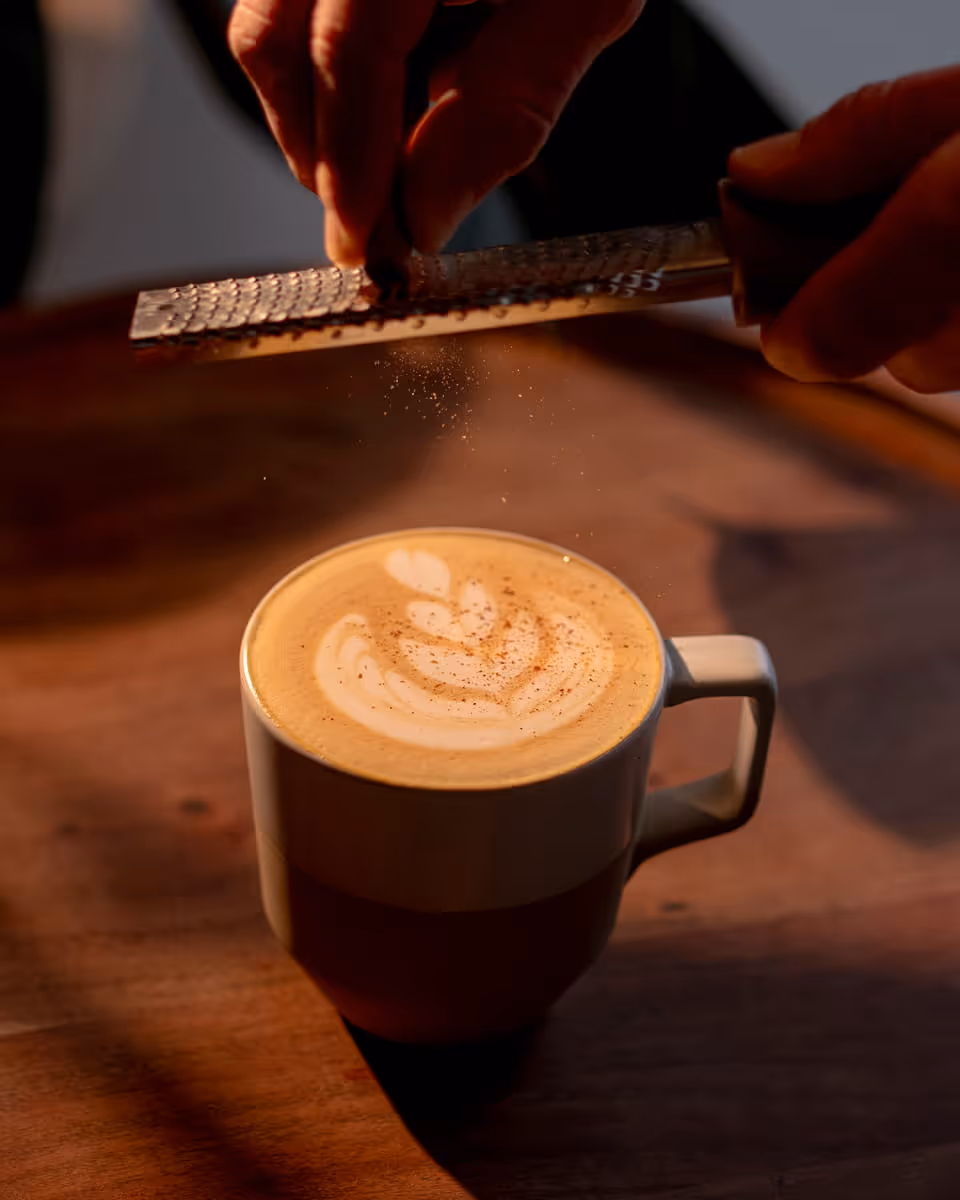 Barista grating fresh spice over a latte with detailed foam art, bathed in warm golden light on a wooden surface.