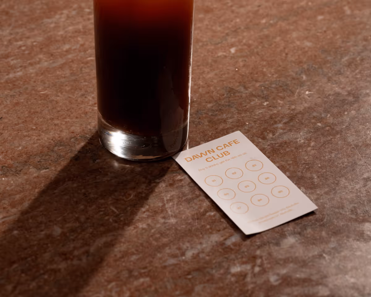 A tall glass of iced black coffee sits beside a Dawn Cafe Club loyalty punch card on a marble countertop in warm light.