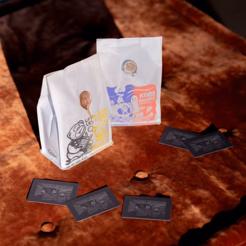 Two specialty coffee bags labeled Costa Rica Las Lajas and Kenya Peaberry beside dark illustrated cards on a tufted leather