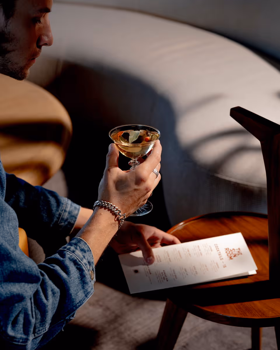 A man in denim holds a martini garnished with a bay leaf while browsing The Sun Room cocktail menu on a sofa.