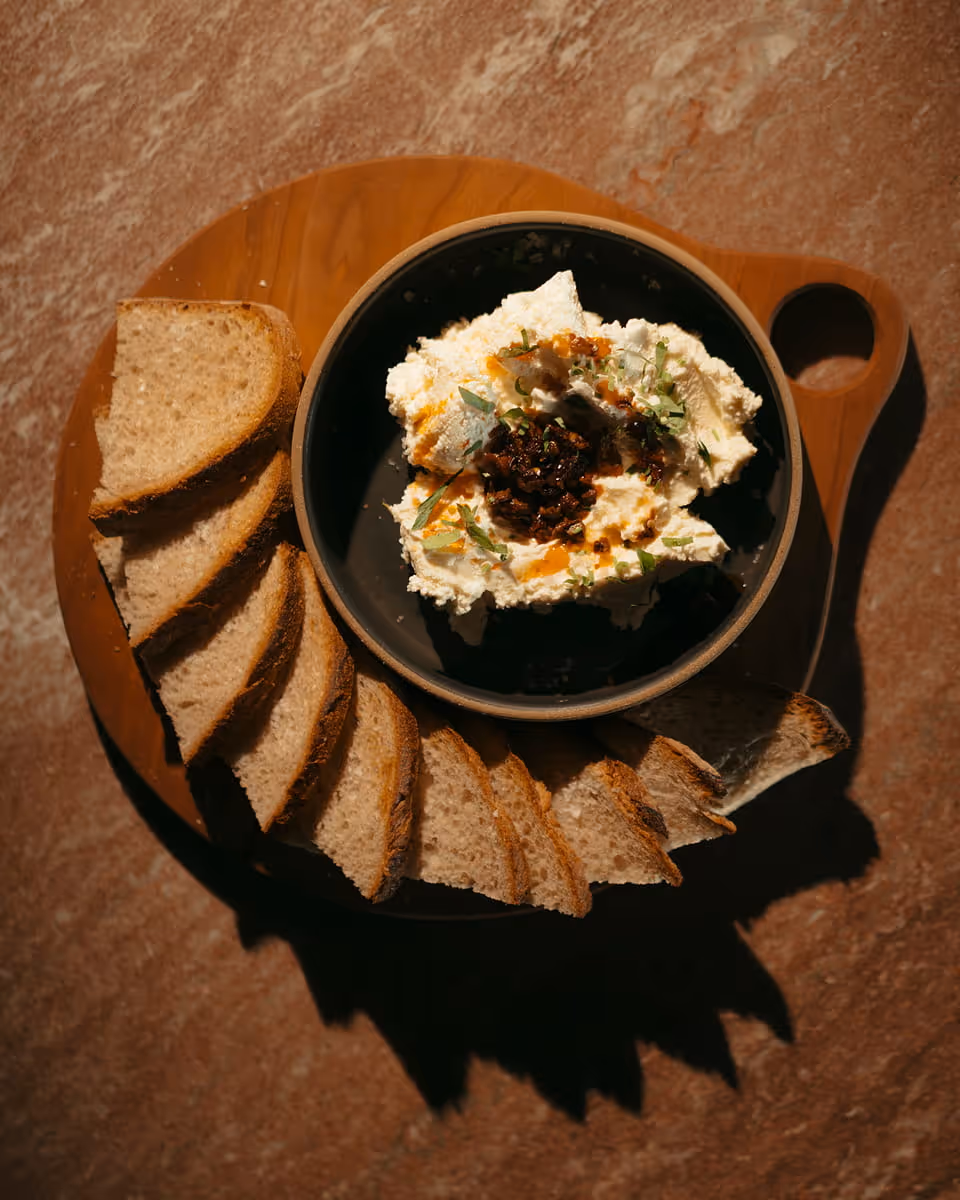 Whipped ricotta dip topped with chili crisp and herbs, served in a dark bowl with toasted bread slices on a wooden board.