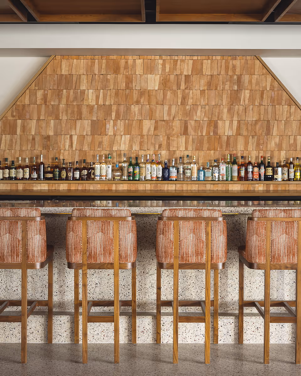 A stylish bar with wooden shingle backdrop, terrazzo counter, rows of spirit bottles, and woven upholstered bar stools.
