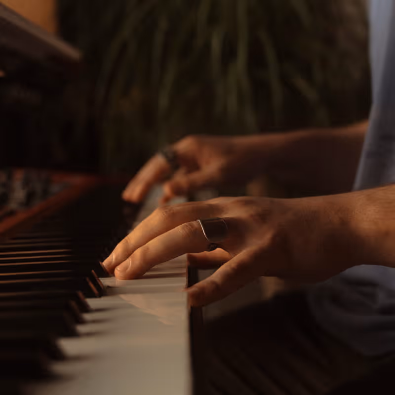 Close-up of hands wearing rings playing piano keys in a warmly lit, intimate musical setting with soft focus background.
