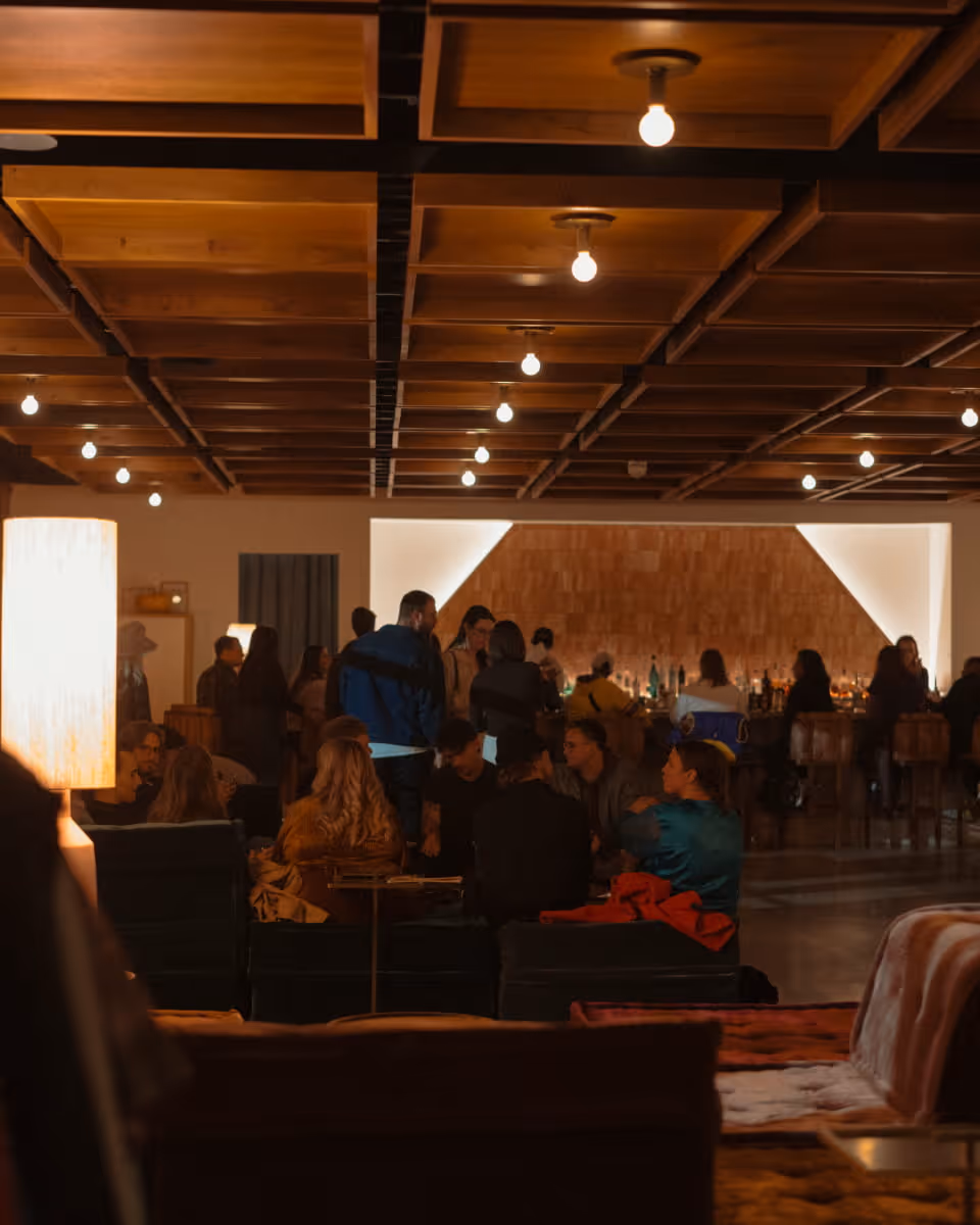 A warmly lit lounge bar with wooden coffered ceiling, globe lights, and groups of people socializing on couches and near