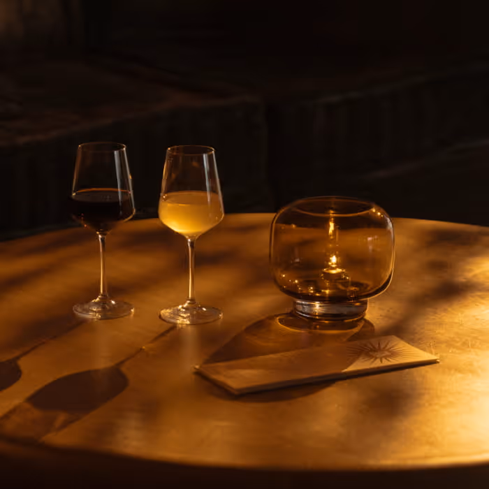 Two wine glasses and an amber glass candle holder on a warmly lit round wooden table in a dimly lit lounge
