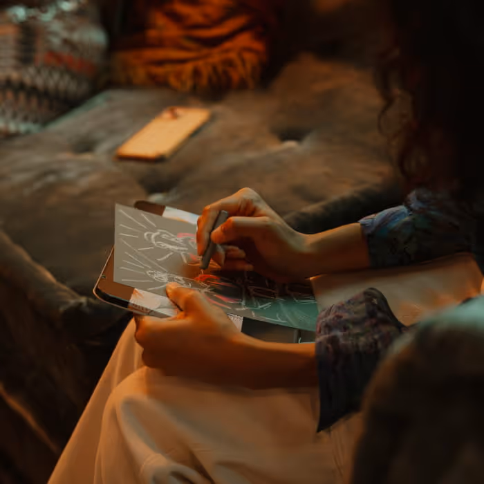 Person sketching a colorful illustration in a small notebook while sitting on a couch in warm ambient lighting