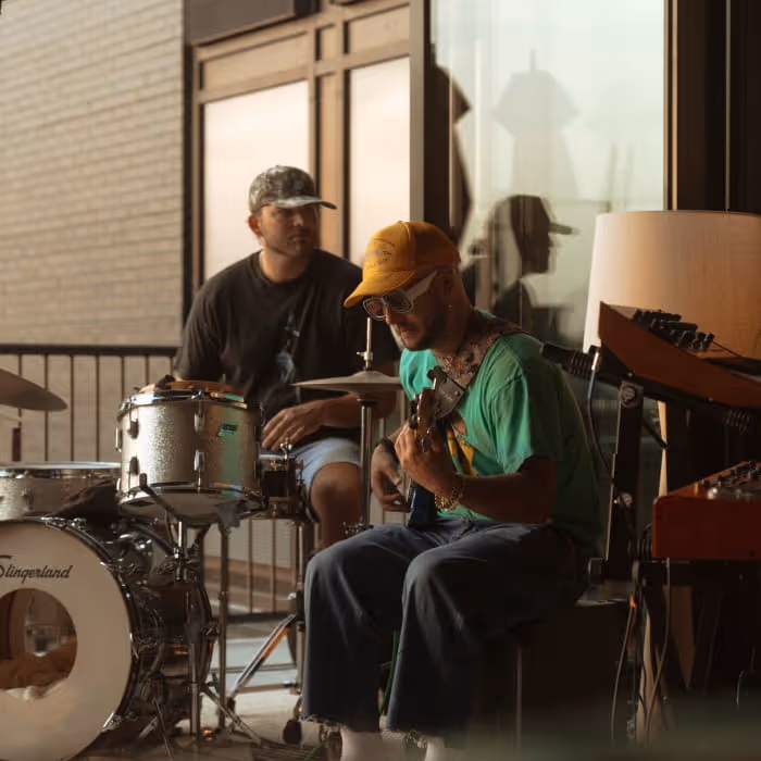 Two musicians jamming outdoors with a Slingerland drum kit and guitar, both wearing caps in golden hour light