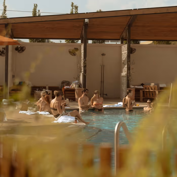 A group of friends relaxing by a sunlit hotel pool under a wooden pergola on a warm summer day
