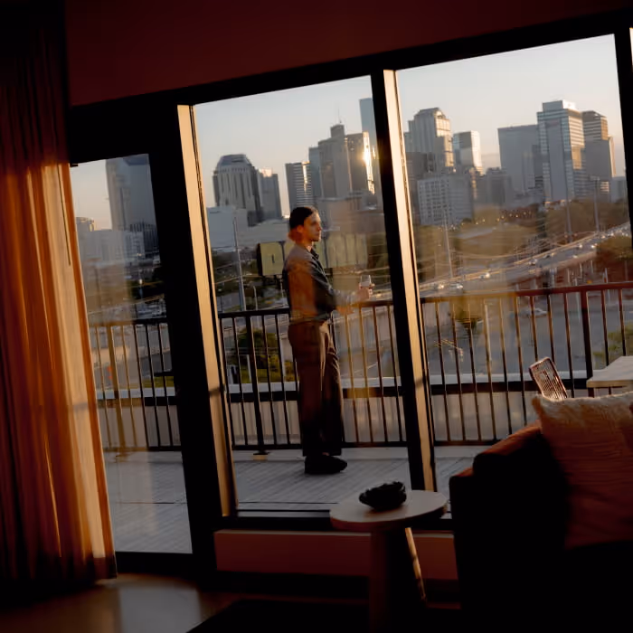 A person stands on a hotel balcony at sunset holding a drink, overlooking a downtown city skyline through floor-to-ceiling