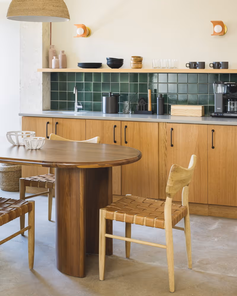Mid-century modern kitchen with green tile backsplash, oak cabinetry, a round walnut dining table, and woven leather chairs.
