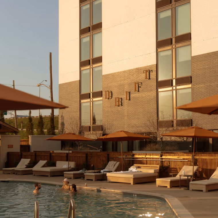 Drift hotel exterior with poolside lounge chairs under orange umbrellas as guests swim in the outdoor pool at golden hour
