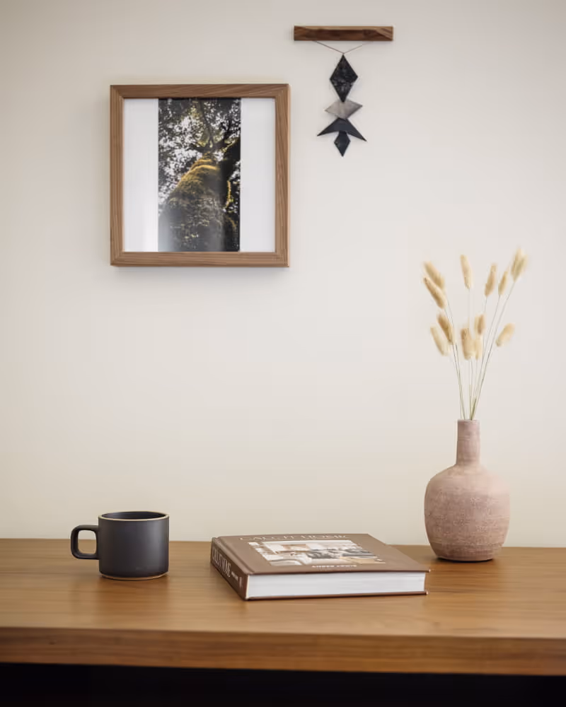 Minimalist hotel desk with a dark ceramic mug, a hardcover book, and a textured vase holding dried bunny tail grasses