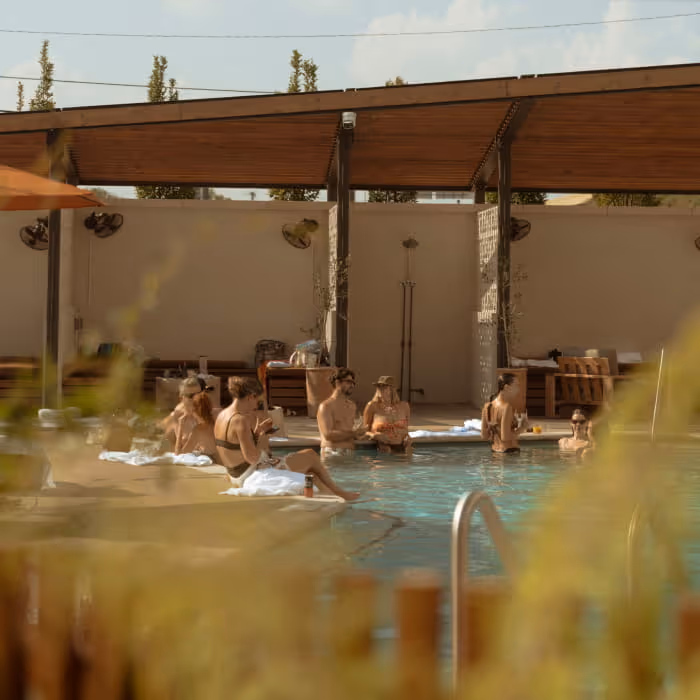 Guests lounging and socializing poolside at a boutique hotel with a wooden pergola and misting fans on a sunny day