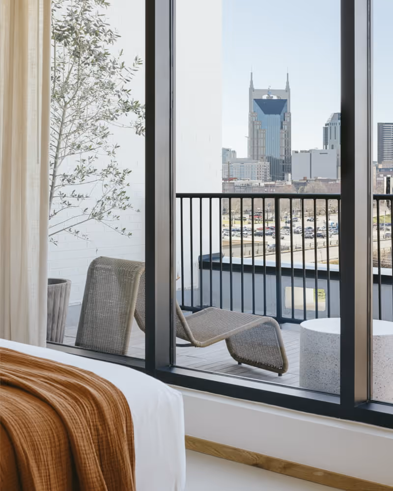 Hotel bedroom view through floor-to-ceiling windows showing a balcony lounge chair overlooking the Nashville downtown