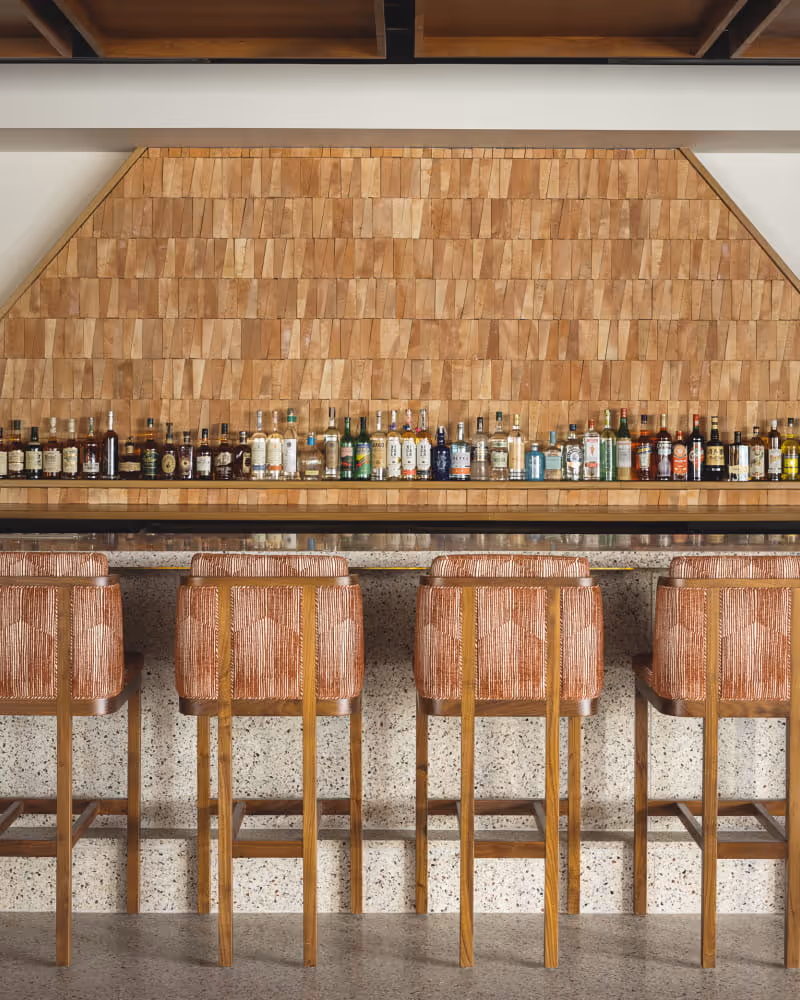 Stylish bar with woven-back stools, terrazzo countertop, and rows of liquor bottles displayed against a wooden shingle wall.