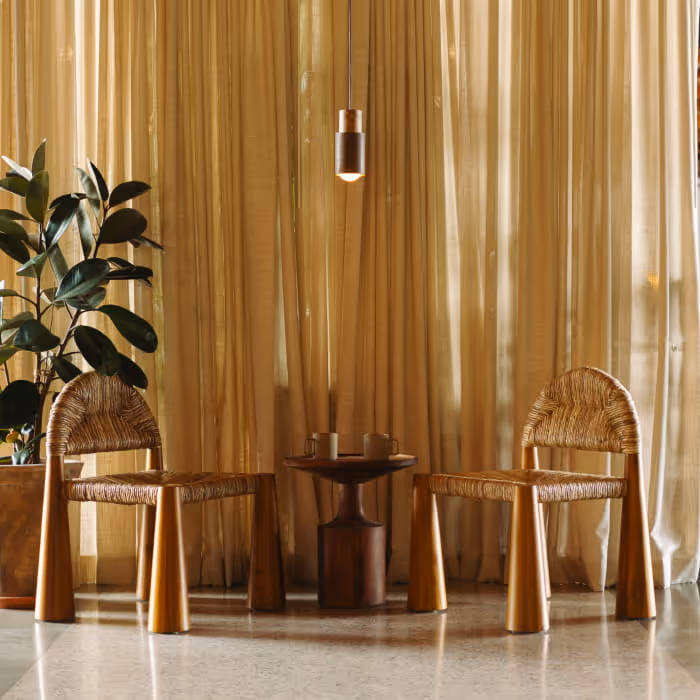 Two woven rattan chairs flank a small wooden side table beneath a pendant light, set against flowing golden sheer curtains.