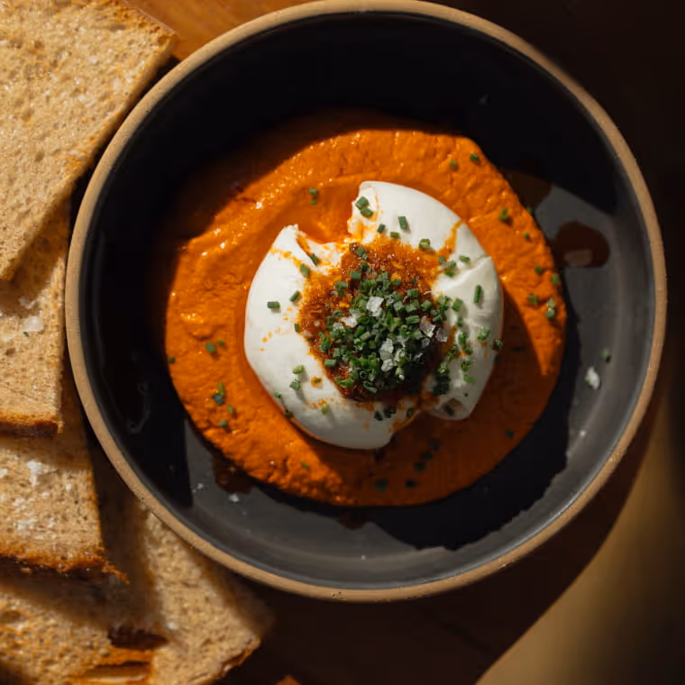 Bowl of vibrant roasted red pepper dip topped with burrata, chives, and chili oil, served alongside sliced flatbread.