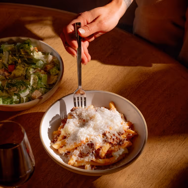 A hand holding a fork digs into a bowl of penne pasta topped with grated Parmesan, alongside a fresh salad and red wine.