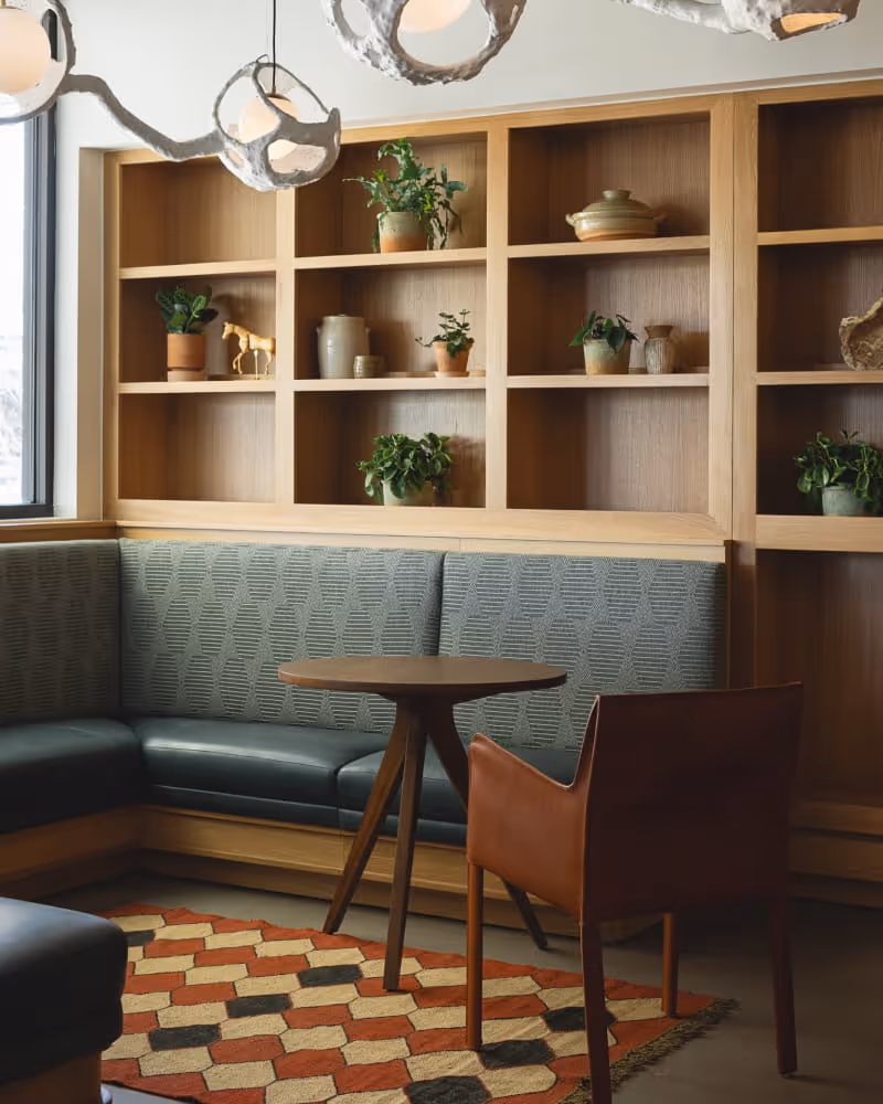 Cozy banquette seating area with wooden shelving displaying potted plants, a round table, leather chair, and geometric rug.