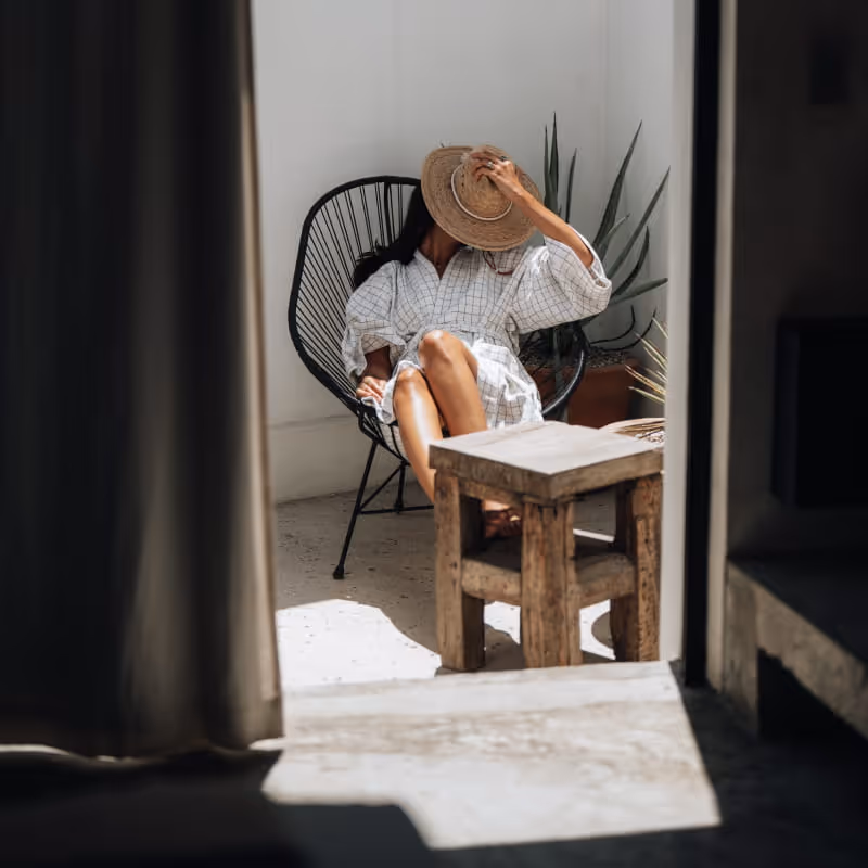 Woman relaxing in a black Acapulco chair on a sunlit patio, holding a straw hat over her face near a wooden table