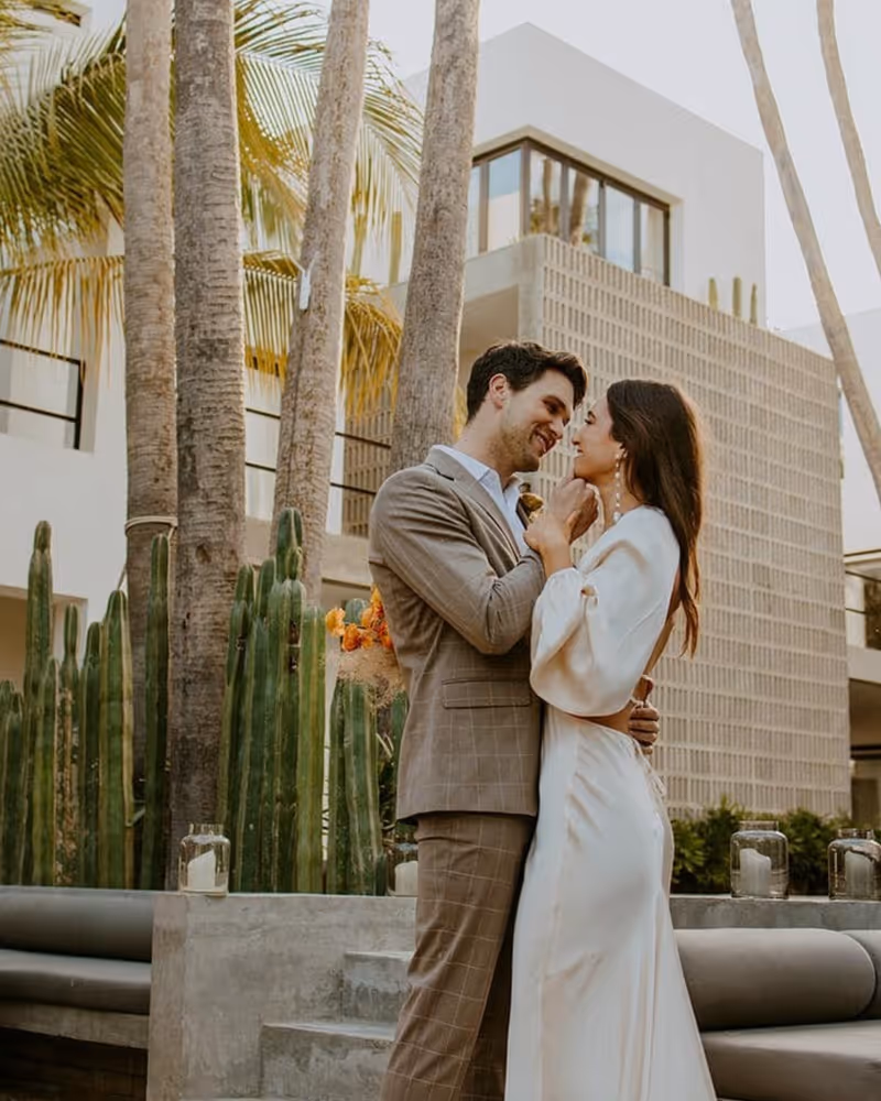 A couple embraces romantically at a tropical wedding venue surrounded by palm trees, cacti, and modern architecture.