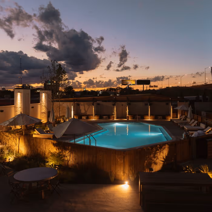 Hotel pool area at dusk with lounge chairs, wooden fence, a picnic bench, and patio seating surrounded by landscaping