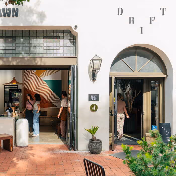 The white facade of Drift Hotel featuring an arched entryway, colorful interior mural, and guests entering the cafe area.